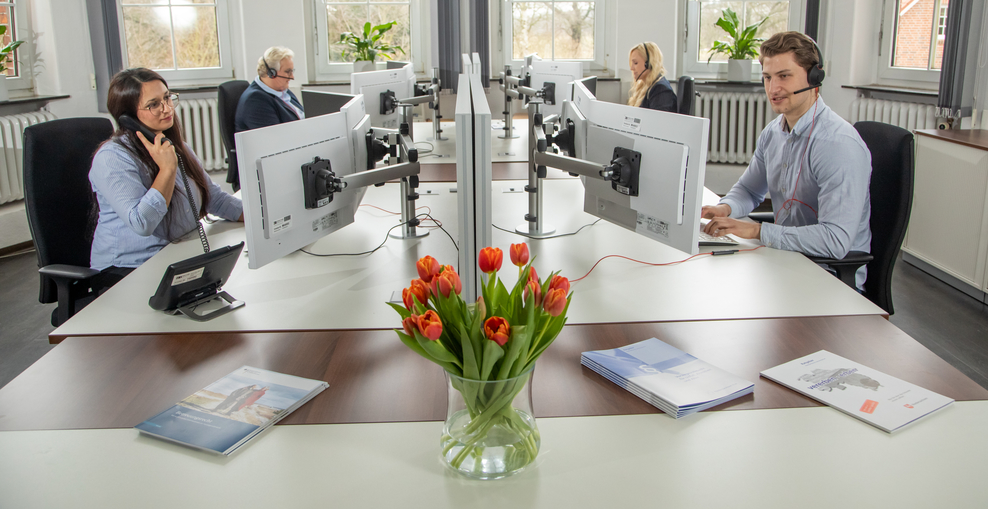 vier Personen sitzen im Büro an ihren Schreibtischen und telefonieren